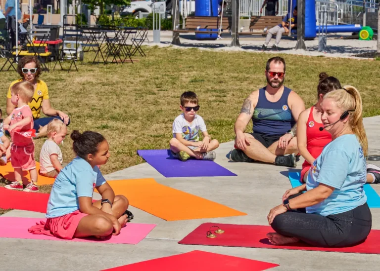 Eight people, adults and children, are seen doing yoga outside on a sunny day.