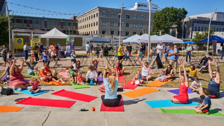 A dozen people are seen doing yoga together outside on a sunny day.