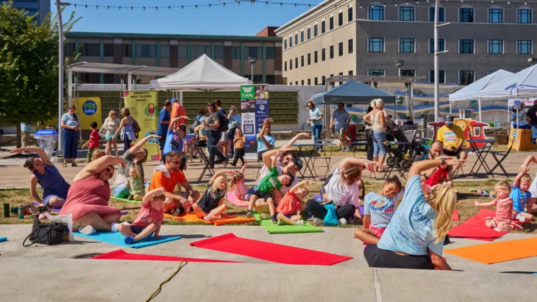 Several children with their parents can be seen doing yoga on yoga mats outside on a sunny day.