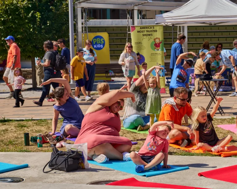More than a dozen people are visible. Children and their parents can be seen doing yoga outside on a sunny day.