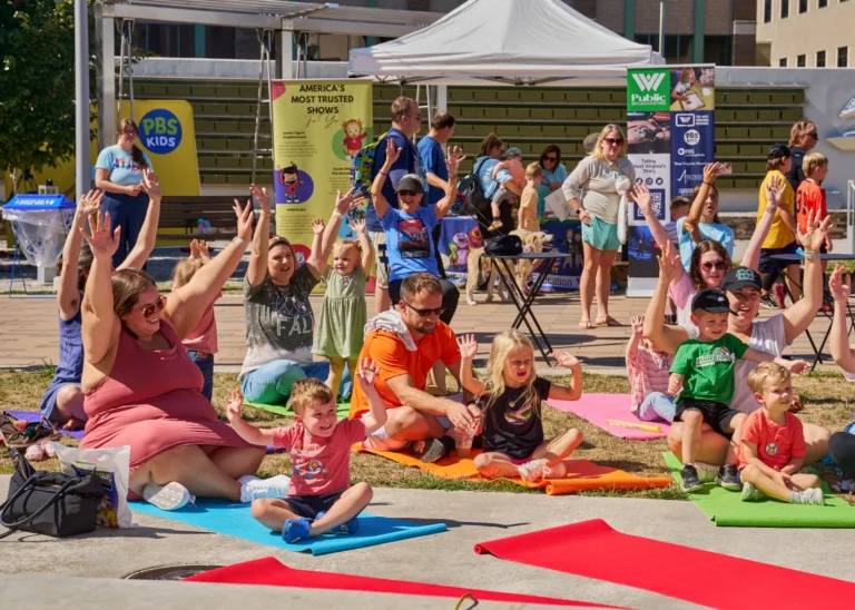 More than a dozen people are visible. Children and their parents can be seen doing yoga outside on a sunny day.