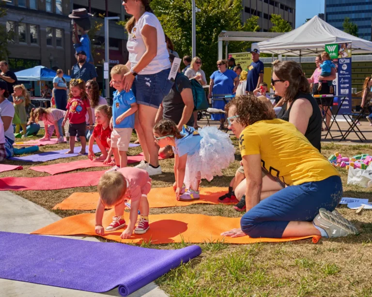 A dozen people are shown doing yoga outside on a sunny day. The people range in age from adult to young child.