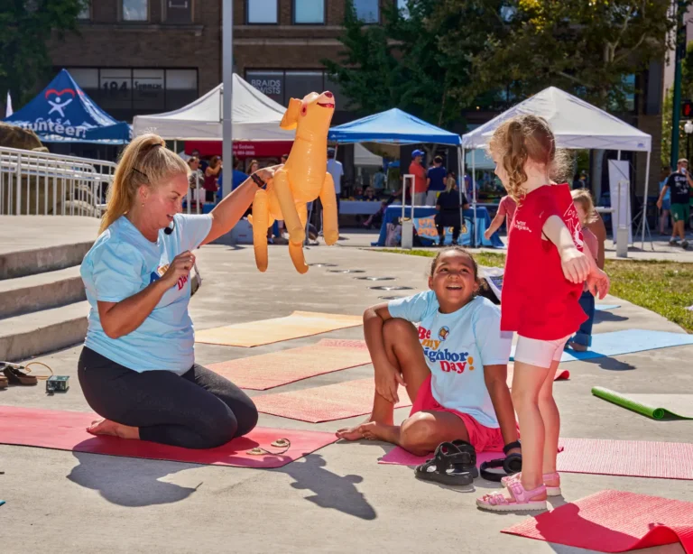 Two children smile and have fun while doing yoga. A yoga teacher holds up a balloon dog and smiles at them.