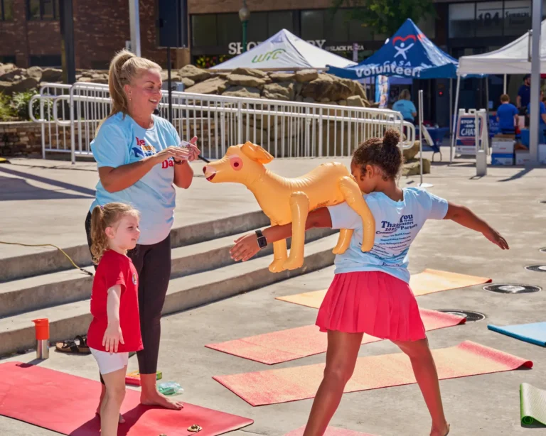 A young girl does yoga while balancing a balloon dog on her shoulder.
