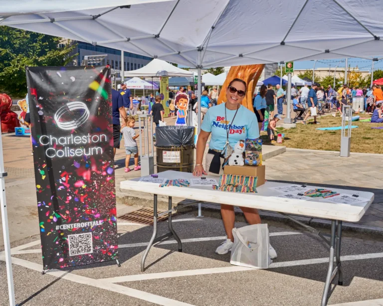 A woman stands at a table and smiles for the camera. Next to hear is a popup banner that reads, "Charleston Coliseum."