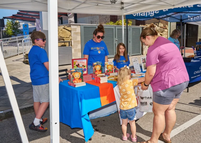 Two adults, and three children, stand at a vendor table where several books are stacked.