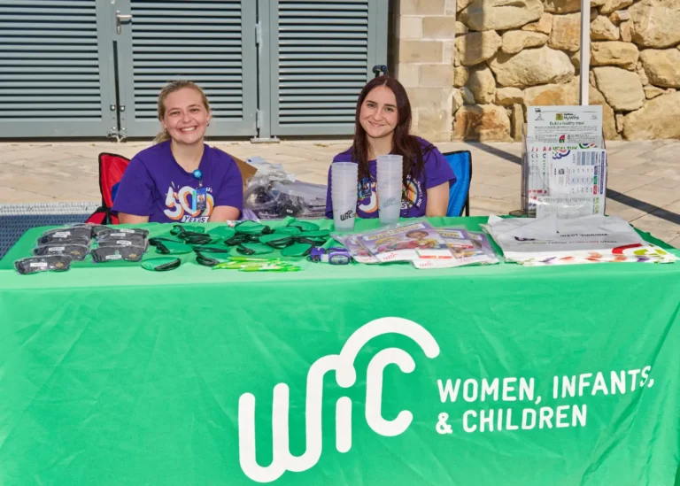 Two women sit behind a vendor table and smile for the camera. There is a tablecloth with text that reads, "WIC, women, infants, and children."
