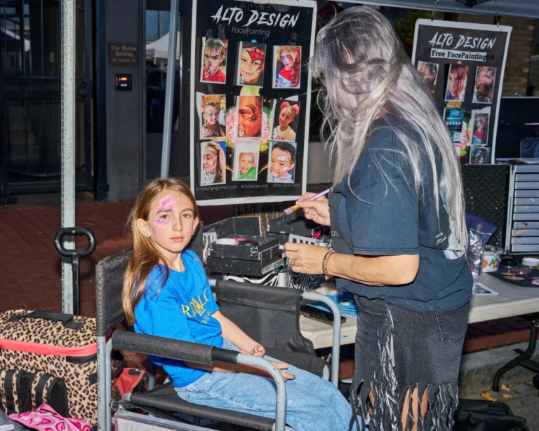 A young girl is getting her face painted. She sits in a chair as a woman with long, gray hair gets ready to paint the girl's face.