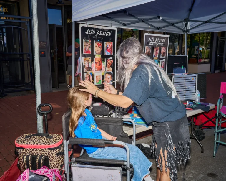A young girl is getting her face painted. She sits in a chair as a woman with long, gray hair gets ready to paint the girl's face.