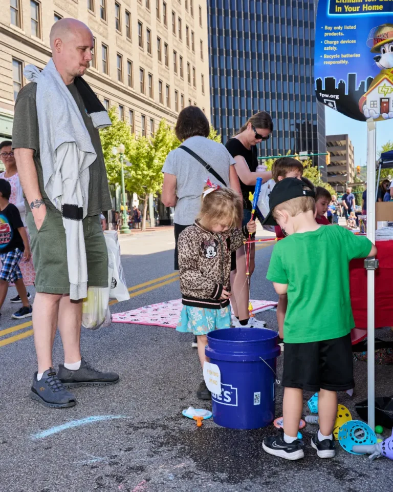 An adult man stands near two small children who stand around a bucket.
