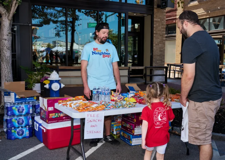A man stands at a vendor table. A sign on the table reads, "Free snacks and drinks." An adult man and a small girl stand at the table.