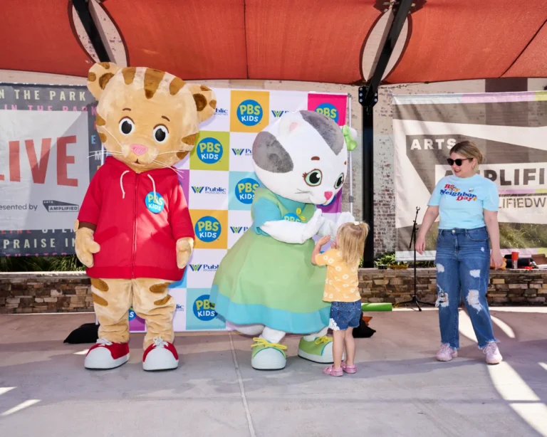 A little girl gives Katerina Kittycat a high five. An adult smiles and watches from the side. Daniel Tiger is seen looking toward the camera.
