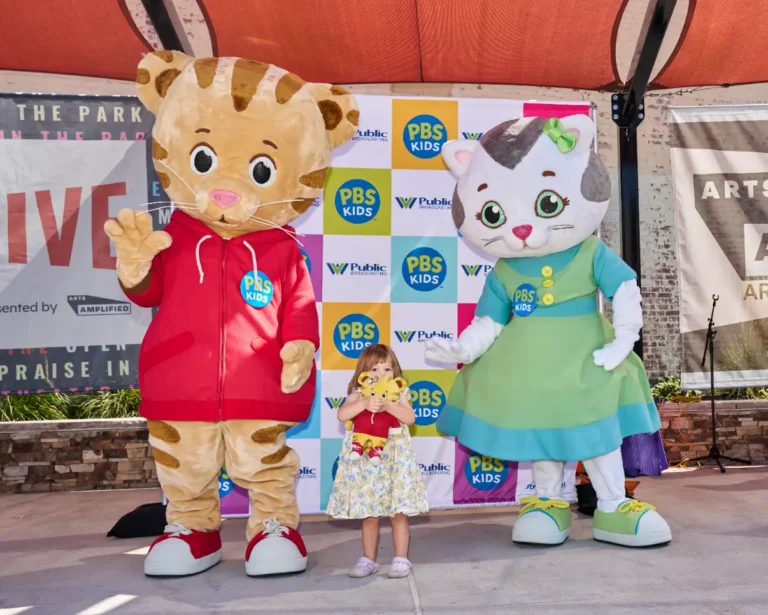Daniel Tiger and Katerina Kittycat with a small girl. The girl is squeezing a Daniel Tiger stuffed toy.