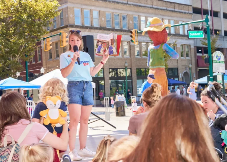 A woman looks down toward people listening to her read a book aloud outside on a sunny day.