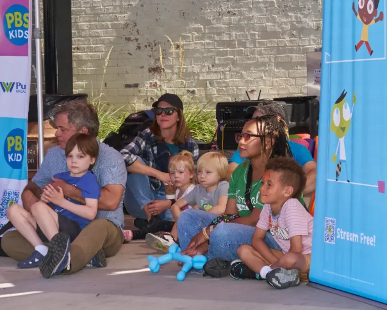 Several people, adults and children, sit on the ground and listen to a storybook read aloud.