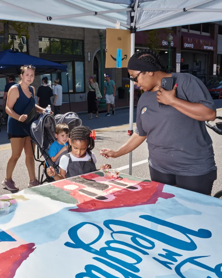 A woman helps a girl paint a mural. Behind them is a mother and a child in a stroller.
