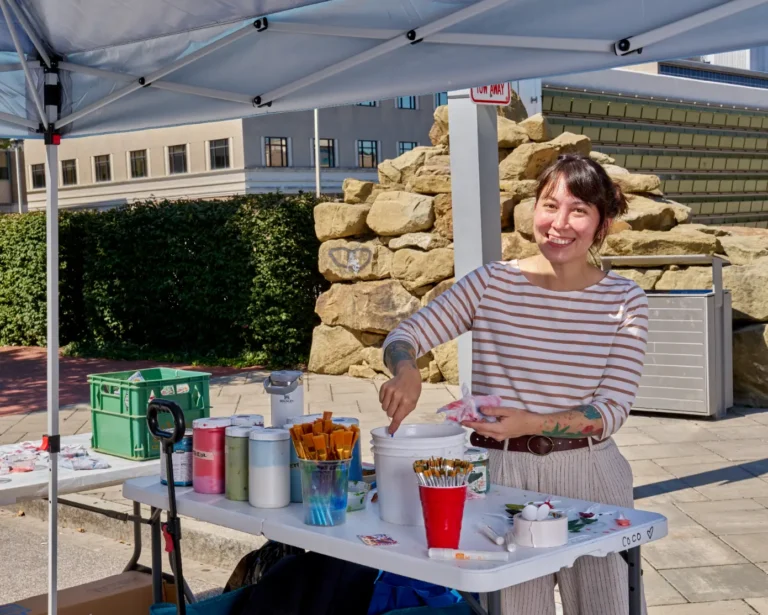 A woman looks toward the camera and smiles. She is standing at a table with paint and dozens of paint brushes.