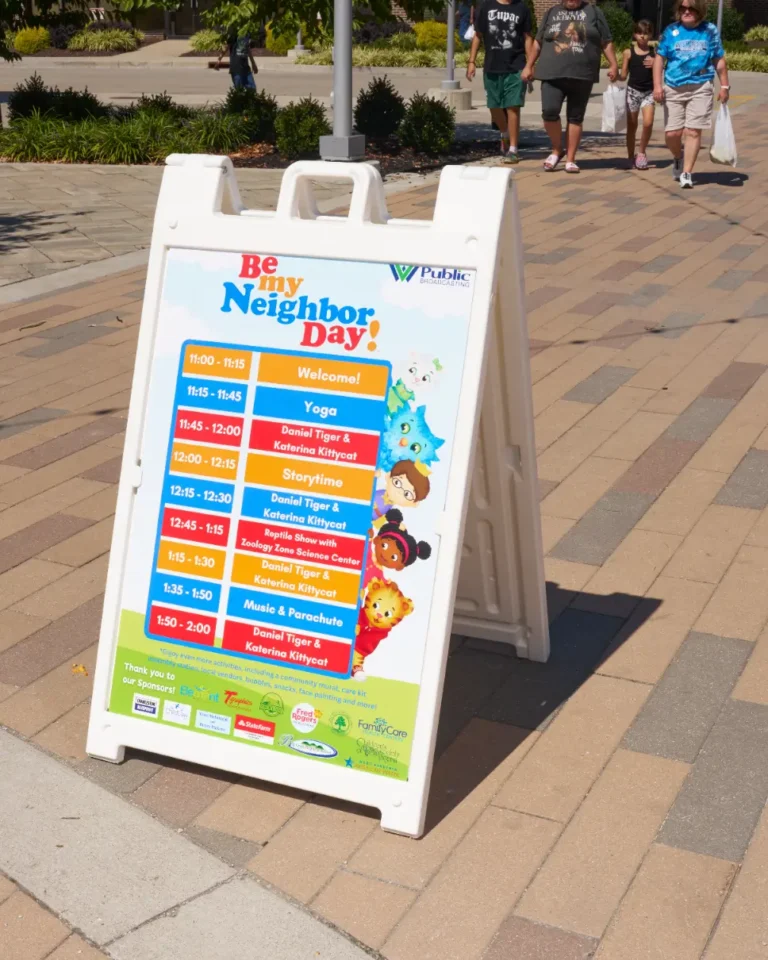 A close up photo of a standup poster that says, Be My Neighbor Day! There is a schedule of events for the day.
