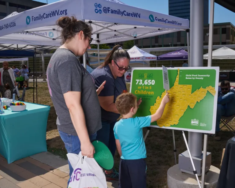 Two adults and one child. They stand in front of a poster of the WV state.