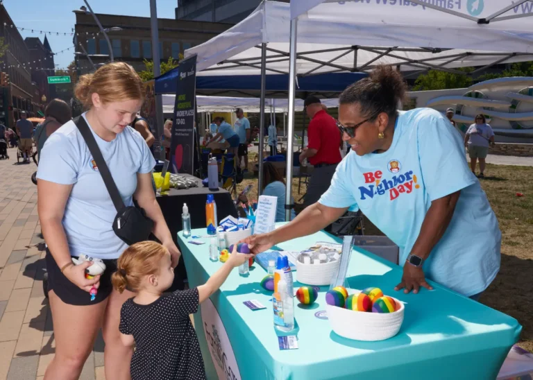 A woman gives a small girl an item off a vendor table. Next to the girl is an older girl.