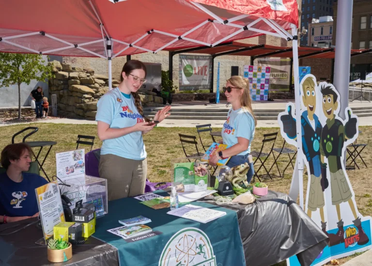 Two adults stand behind a vendor table and speak to each other, smiling.