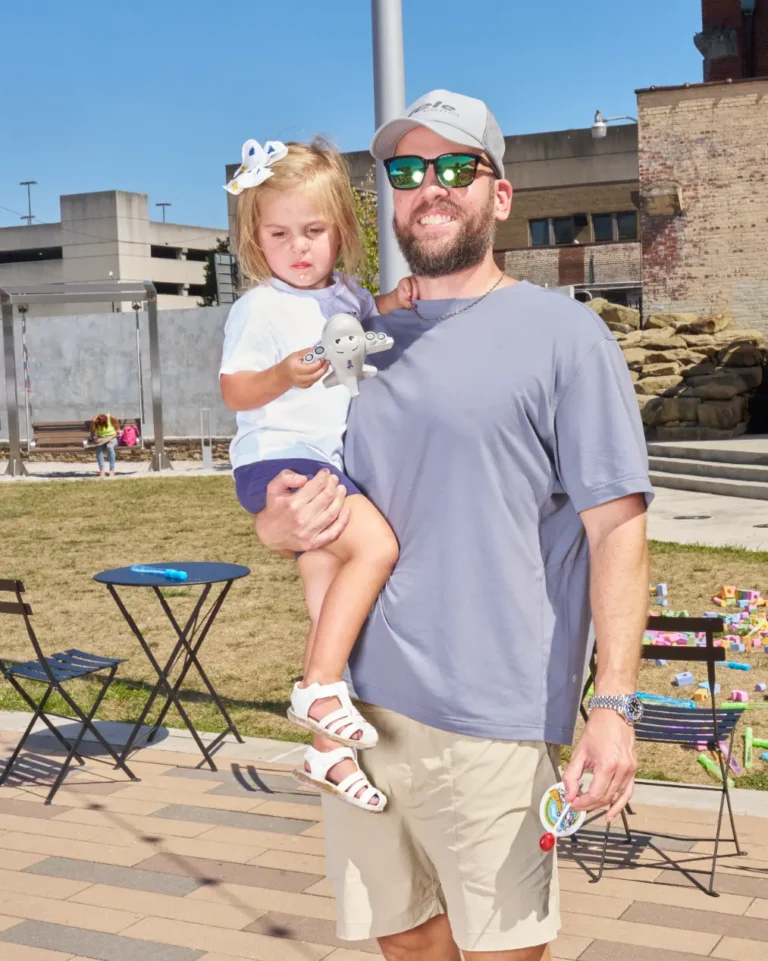 A man wearing sunglasses and a ball cap holds a young girl in his arms. The girl is looking off camera and holding a small toy.