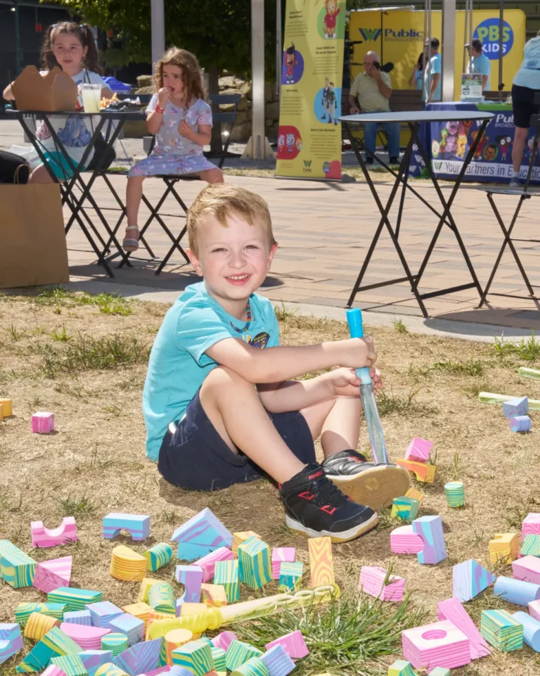A young boy playing with blocks on the ground looks at the camera and smiles.