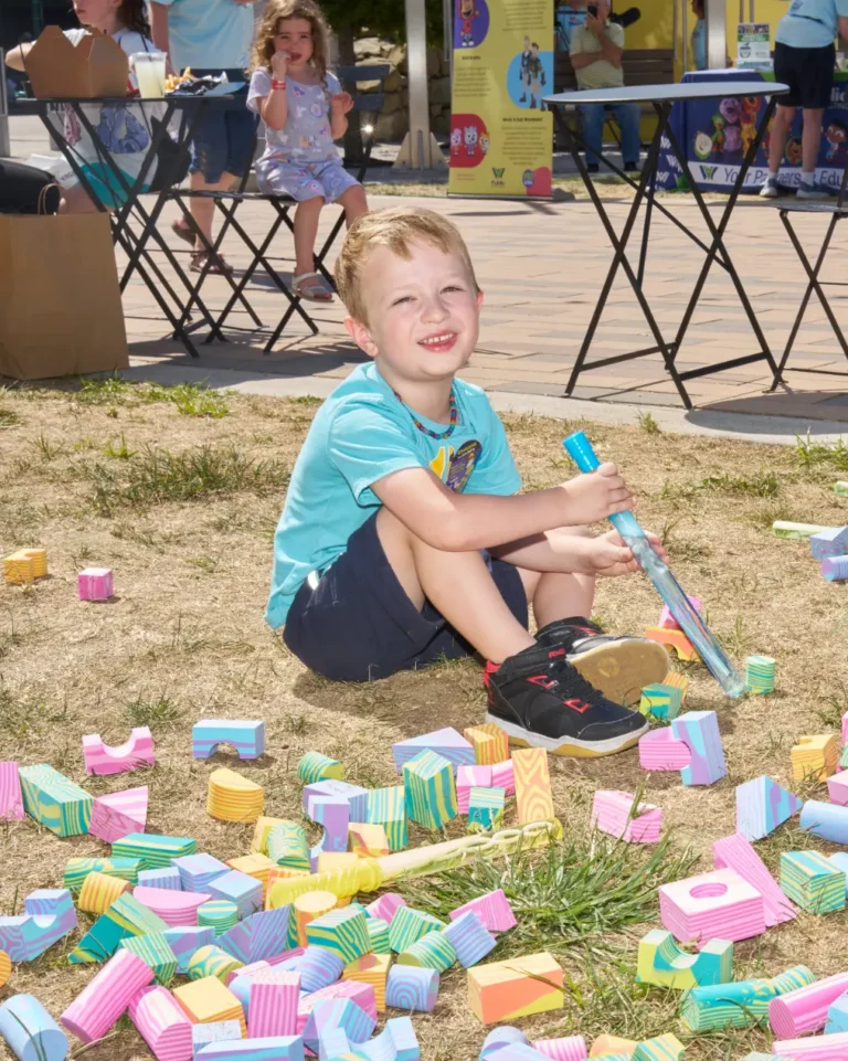 A young boy playing with blocks on the ground looks at the camera and smiles.