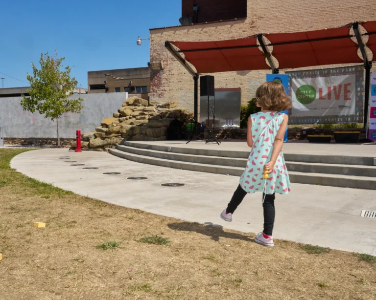 A young girl wearing a blue and pink dress walks away from the camera on a sunny day.