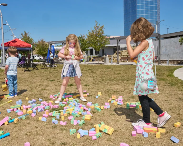 Two children play with blocks outside on a sunny day. The blocks are a colorful pastel color.
