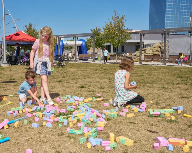 Three children play with colorful, pastel blocks outside on a sunny day.