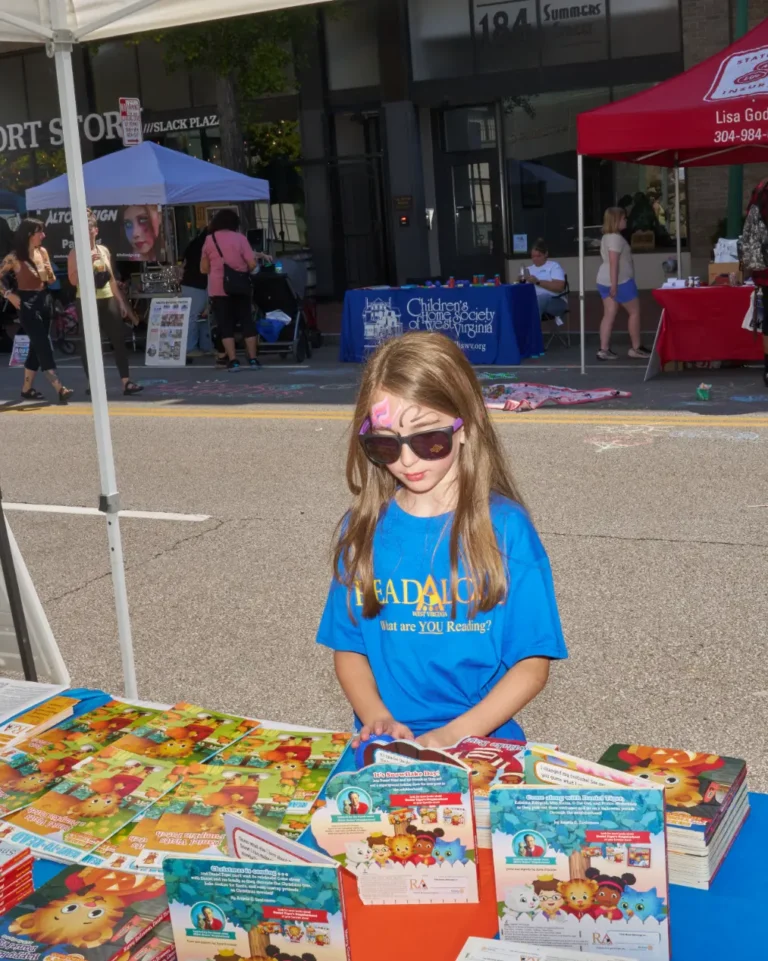A young girl with her face painted and wearing sunglasses looks at several items shown on a table.