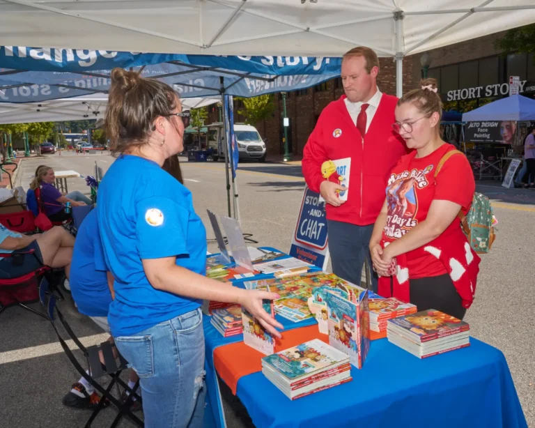 One adult woman speaks to two people at a vendor table. The two people, a man and a woman, wear red and look at the books stacked on the table.