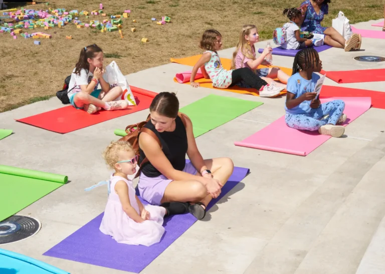 Half a dozen people, two adults and children, sit on yoga mats outside on a sunny day.