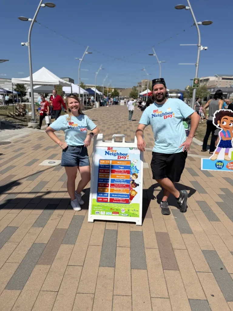 Two adults, one woman and one man, stand next to a sign smiling. The sign says Be My Neighbor Day. The sign has a calendar of events printed on it.