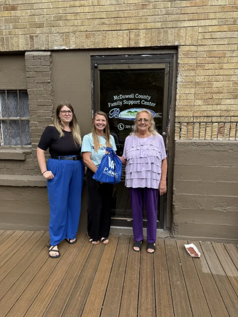 Three adult women stand outside a building. The door behind them reads, "McDowell County Family Support Center." The woman in the center holds a blue WVPB bag.