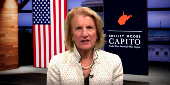 Photo shows a head and shoulder shot of an older woman with short blond hair in a cream colored suit jacket with pearls in front of a backdrop that shows the American flag on one side and an outline of the state of West Virginia with the words "Shelley Moore Capito" on the other