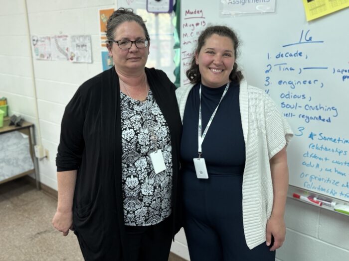 Two women standing next to each other in front of a white board. On the left is Wilfong wearing mostly black with a black and white floral pattern top. The woman on he right is Celi Oliveto who nominated Wilfong. She is wearing a black outfit with a white short sleeved cardigan.