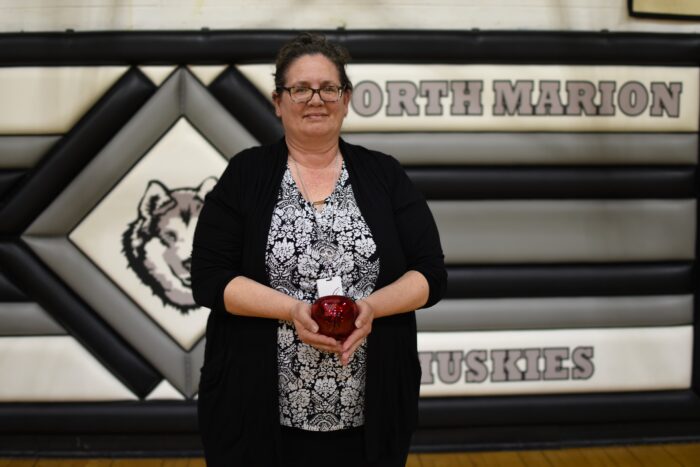 A woman stands in front of striped wall padding that says "North Marion Huskies" with an image of a husky mascot. The woman hold a red, Blenko glass apple paperweight and wears mostly black clothing with a top that is a floral black and white pattern.