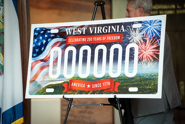 A large poster on an easel shows the new limited-time license plate with an American flag and fireworks in the background and large white sample numbers where a license plate would go.