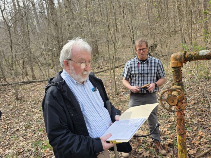 Two men stand in the woods looking at rusty pipes