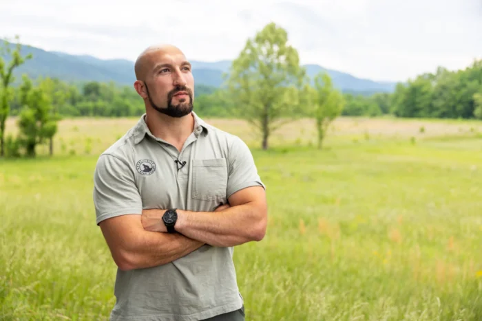 A man in a tan polo looks off camera with his arms crossed over his chest. He is bald and has a trimmed, brown beard and mustache. He stands against a backdrop of grass and trees.