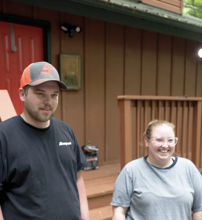 Two young people, a man and a woman, look off camera, smiling. The man is wearing a ball cap, and the woman's hair is pulled up in a bun. She wears glasses.