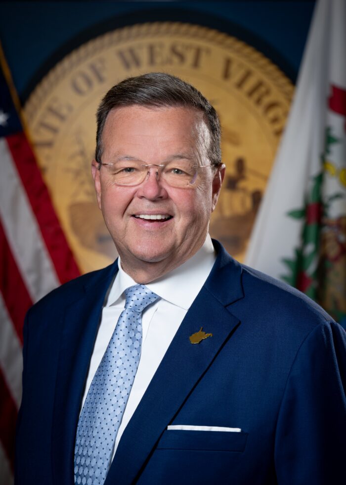 A professional headshot of an older man with brown hair and glasses. The West Virginia state seal is seen behind him, as well as the American flag and the West Virginia flag. He wears a blue blazer, light blue tie, white button up shirt, and has a pin of West Virginia on his lapel.