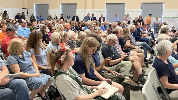 A crowd fills almost every seat in a large room, with more people standing against the far wall of the room.
