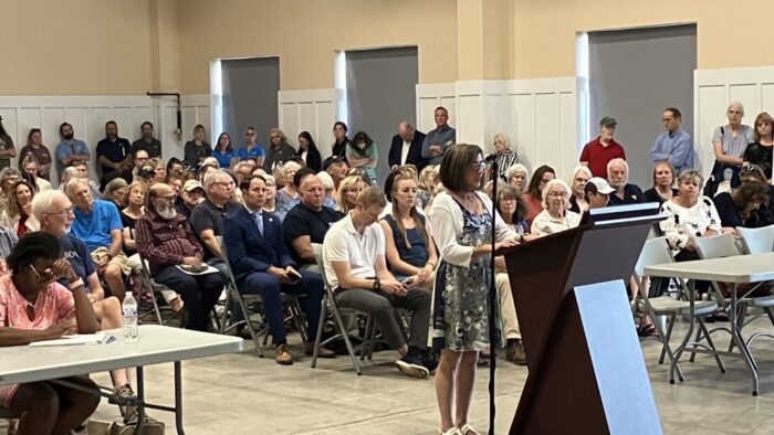 A woman stands at a wooden lectern reading a written statement. Behind her seats are filled with people listening on. More people stand against the walls around the room.