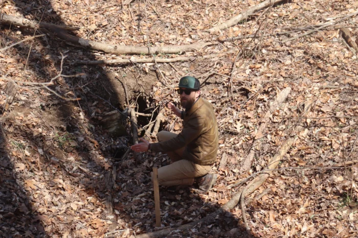 A man crouches in fallen leaves next to a hole in a hill. He wears a ball cap and sunglasses, a brown jacket and tan pants.
