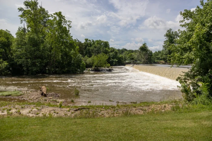A wide shot of a dam affecting the flow of water in a river. There are several green trees in the distance, against a cloudy, blue sky.