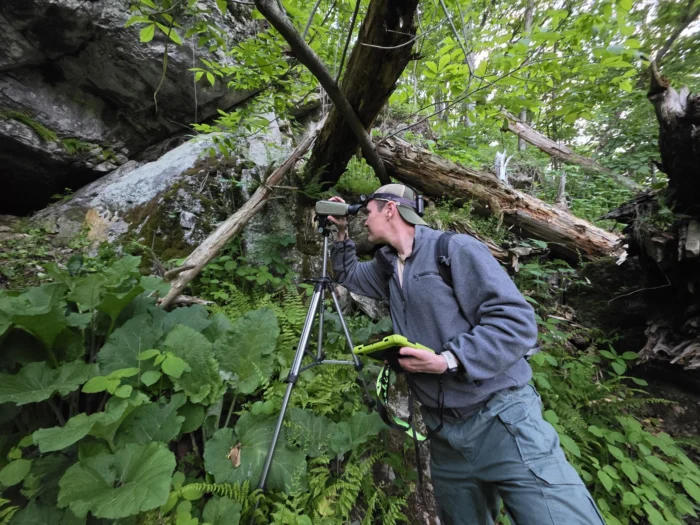 A low angled shot of a man looking into a camera at the mouth of a cave out in a forest. He holds a tablet and wears a ball cap backwards. He is surrounded by green vegetation and tree trunks.