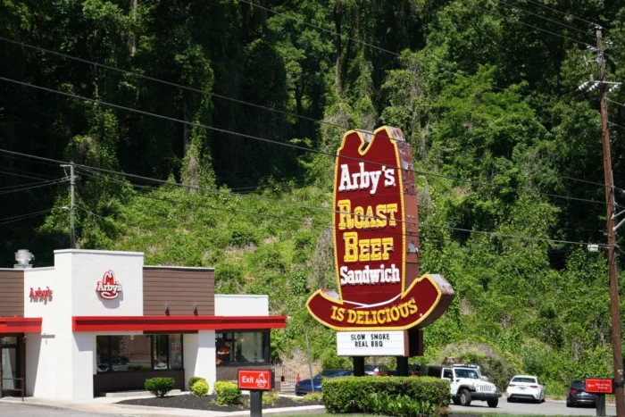A landscape photo of an Arby's sign shaped like a cowboy hat. There is a lot of green vine behind the building. 